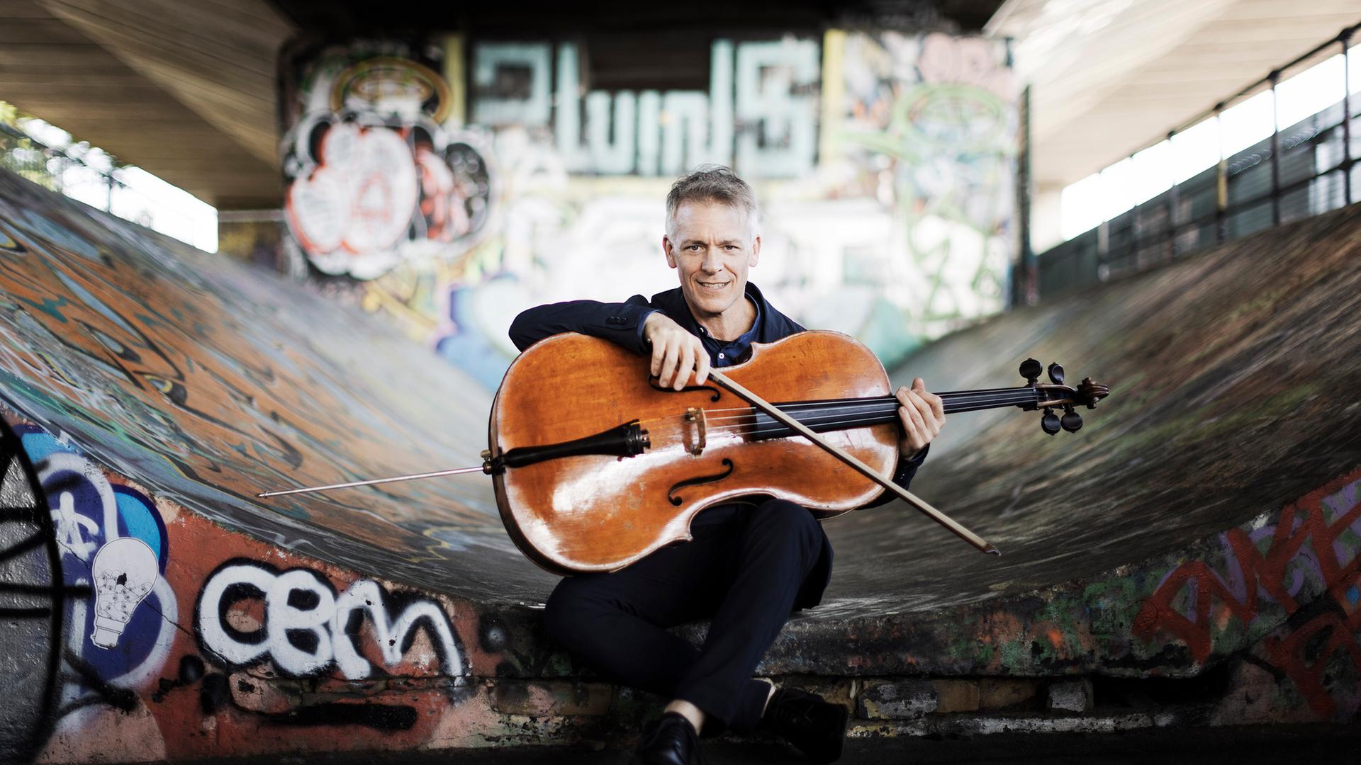 Alban Gerhardt sitzt mit seinem Cello unter einer Brücke, die wilde Graffiti aufweist.