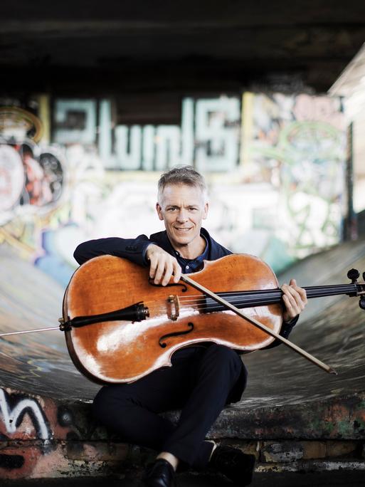 Alban Gerhardt sitzt mit seinem Cello unter einer Brücke, die wilde Graffiti aufweist.