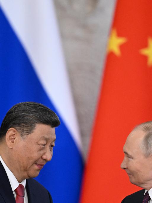 Xi Jinping (links) und Wladimir Putin (rechts im Bild) stehen vor der russischen und der chinesischen Flagge und schütteln sich die Hände. Xi Jinping (links) und Wladimir Putin (rechts im Bild) stehen vor der russischen und der chinesischen Flagge und schütteln sich die Hände.