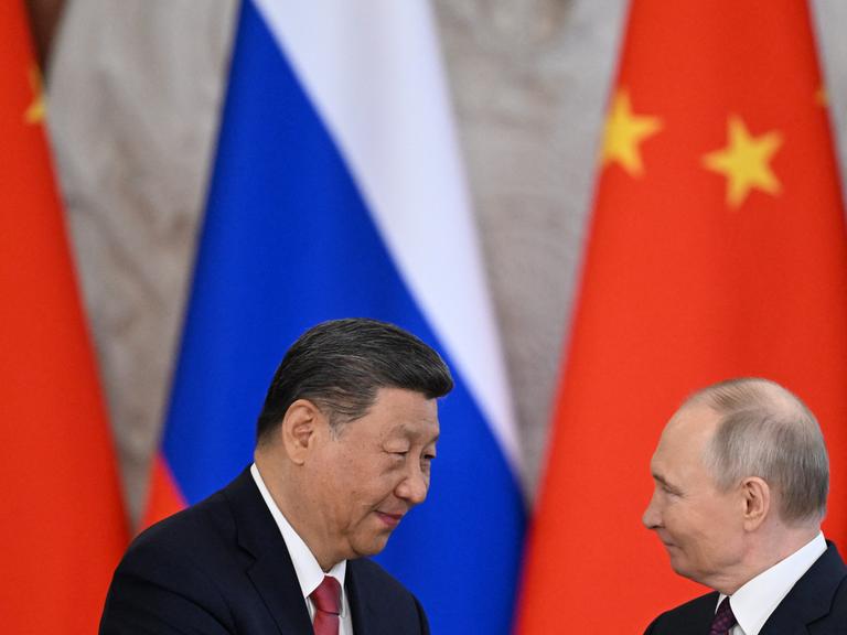 Xi Jinping (links) und Wladimir Putin (rechts im Bild) stehen vor der russischen und der chinesischen Flagge und schütteln sich die Hände.