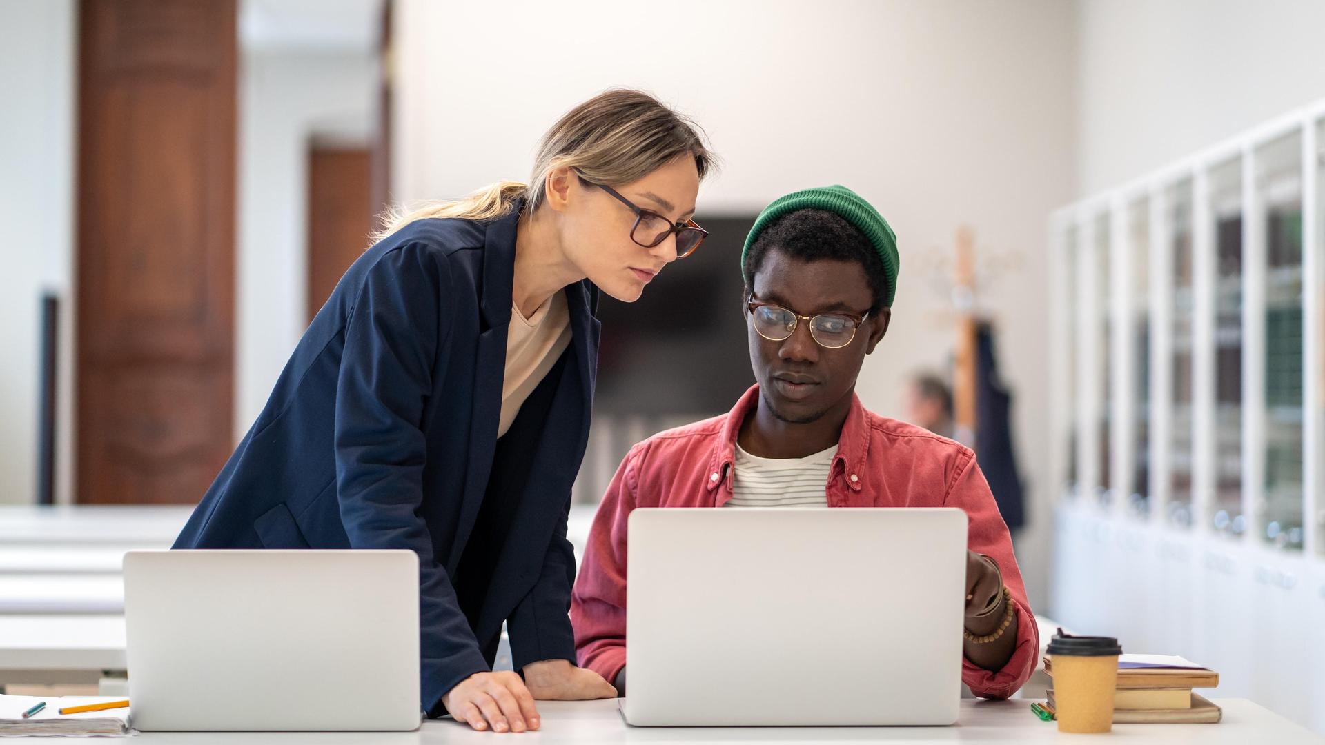 Ein junger Mann sitzt an einem Laptop, eine junge Frau steht ebenfalls an einem Laptop und lehnt sich zu ihm. Er zeigt ihr etwas auf seinem Monitor