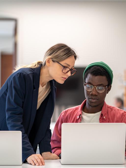 Ein junger Mann sitzt an einem Laptop, eine junge Frau steht ebenfalls an einem Laptop und lehnt sich zu ihm. Er zeigt ihr etwas auf seinem Monitor