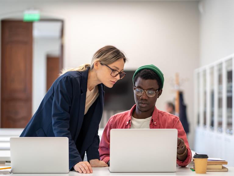 Ein junger Mann sitzt an einem Laptop, eine junge Frau steht ebenfalls an einem Laptop und lehnt sich zu ihm. Er zeigt ihr etwas auf seinem Monitor