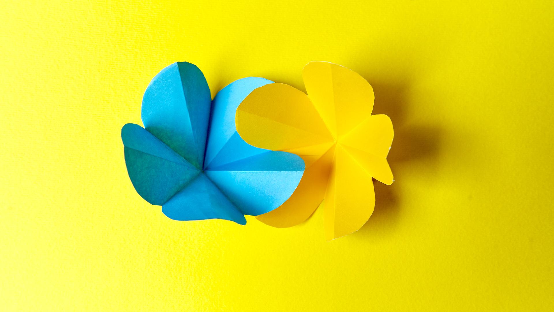 Zwei gefalte Papierblumen sind ineinandergeschoben. Sie sind blau und gelb. Der Hintergrund ist ebenfalls gelb.