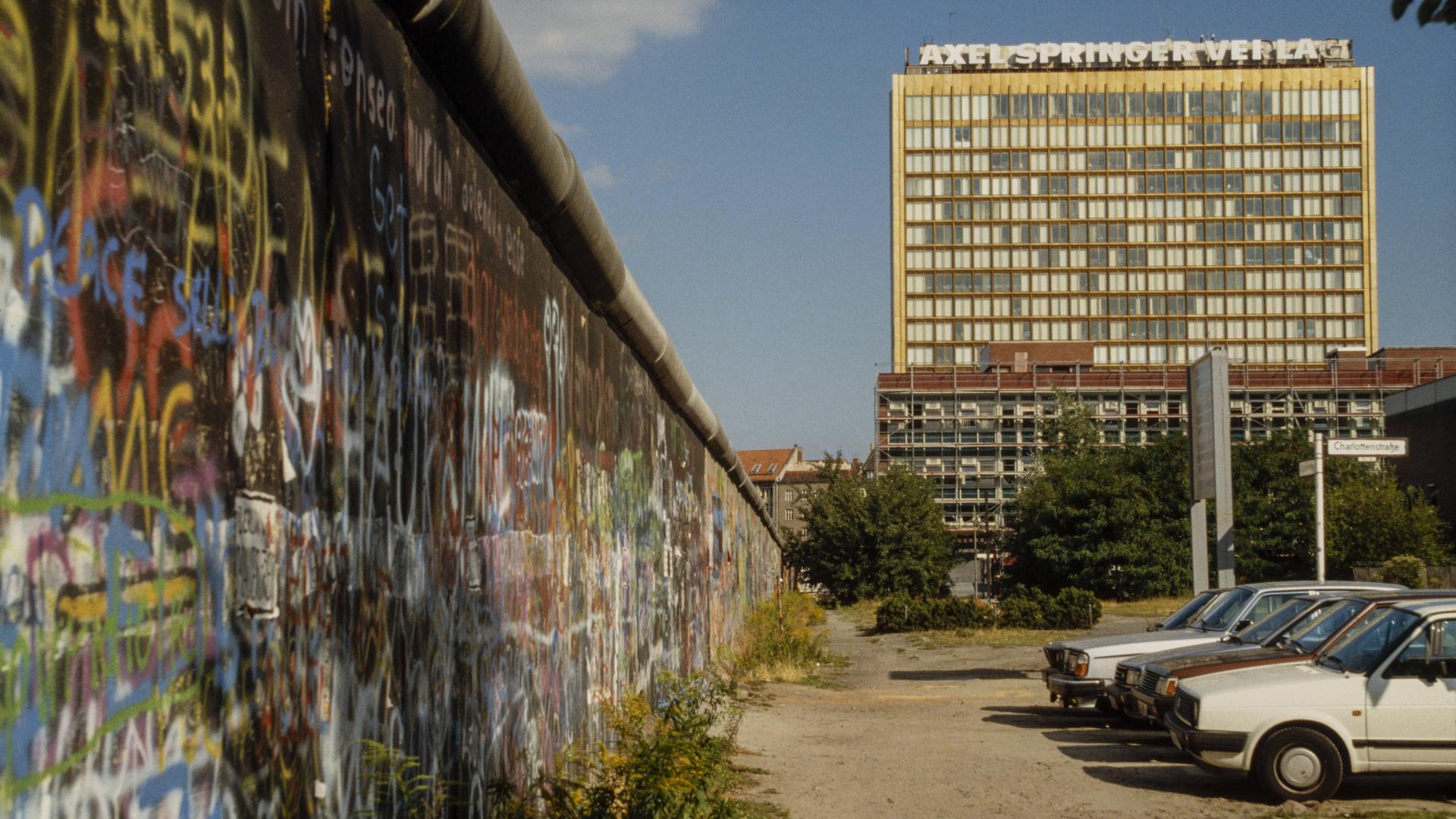 Das Hochhaus des Axel-Springer-Verlags steht hoch neben der bunt bemalten Berliner Mauer. Im Vordergrund parken Autos im typischen Stil der 1980er-Jahre