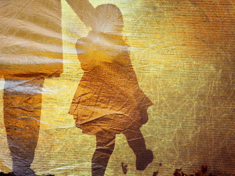 Der Schatten eines kleinen Mädchens an der Hand einer erwachsenen Person bildet sich auf einer goldfarbenen Oberfläche ab.