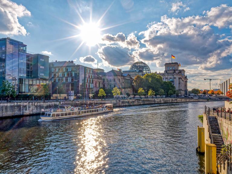 Spreeufer in Herbst mit dem Berliner Reichstag im Hintergrund, die Sonne scheint.
