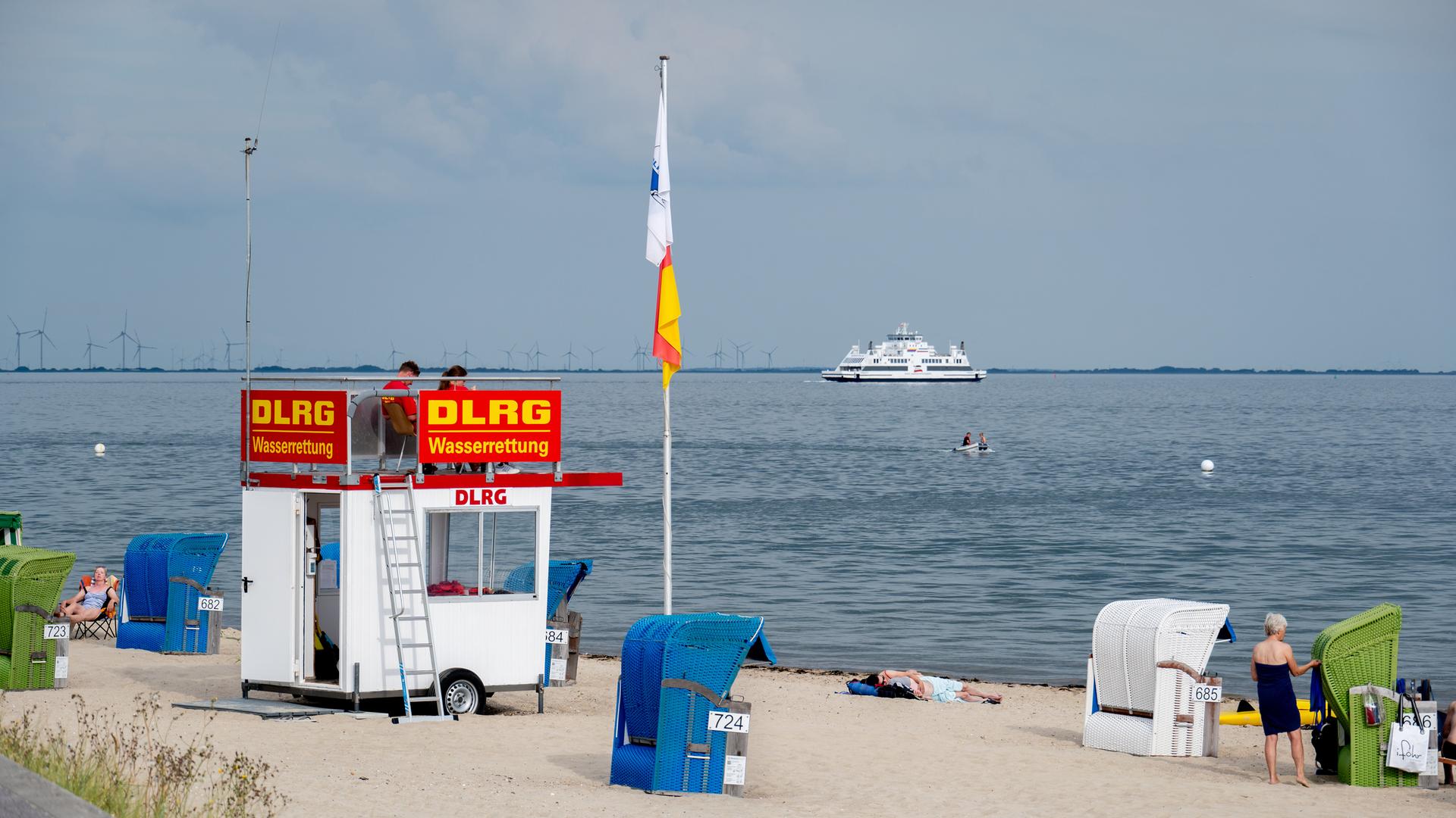 Ehrenamtliche Helfer der DLRG Wasserrettung sitzen auf einem Aufsichtswagen am Strand der Insel. Im Hintergrund ist ein Fährschiff der Wyker Dampfschiffs-Reederei (WDR) im Wattenmeer unterwegs.