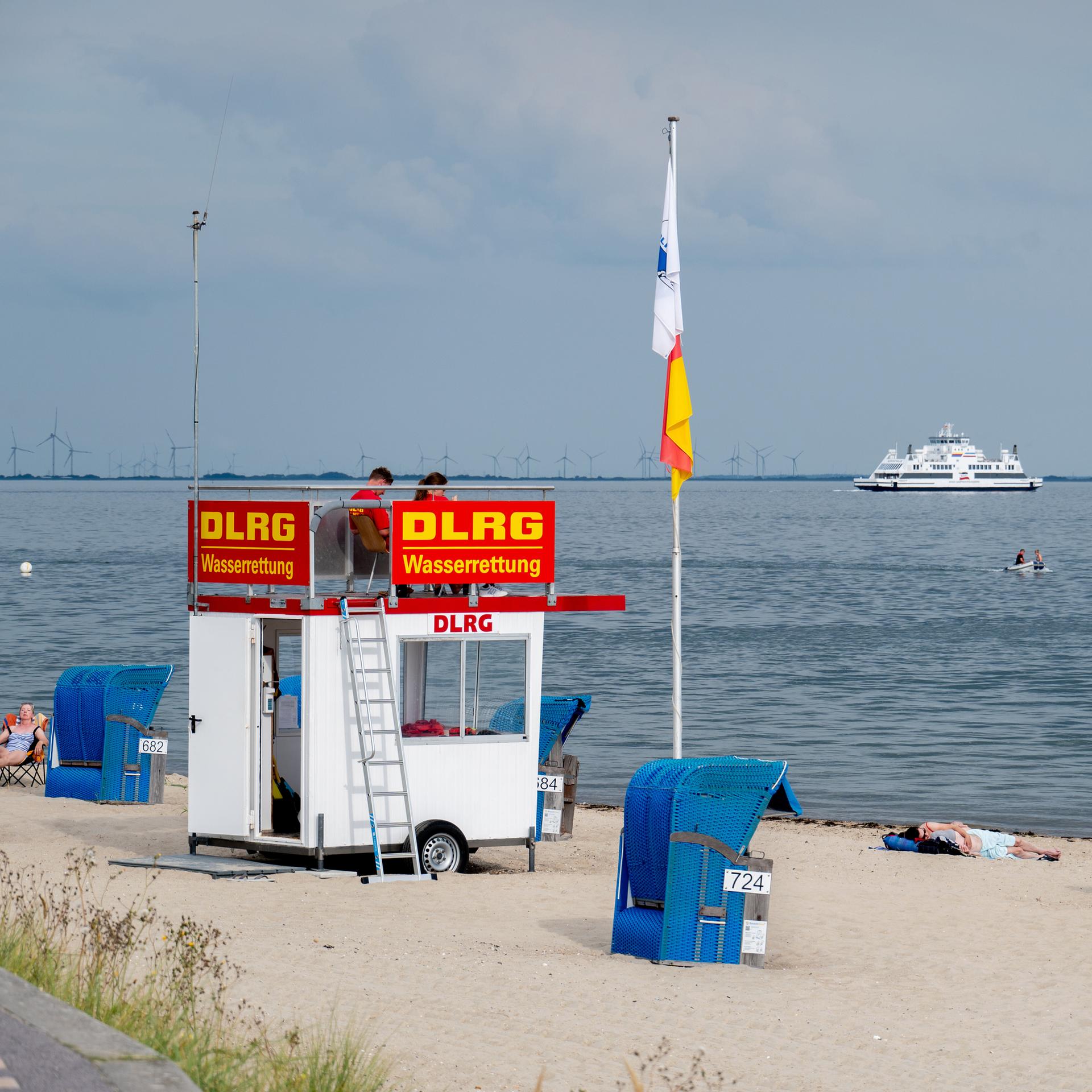 Ehrenamtliche Helfer der DLRG Wasserrettung sitzen auf einem Aufsichtswagen am Strand der Insel. Im Hintergrund ist ein Fährschiff der Wyker Dampfschiffs-Reederei (WDR) im Wattenmeer unterwegs.
