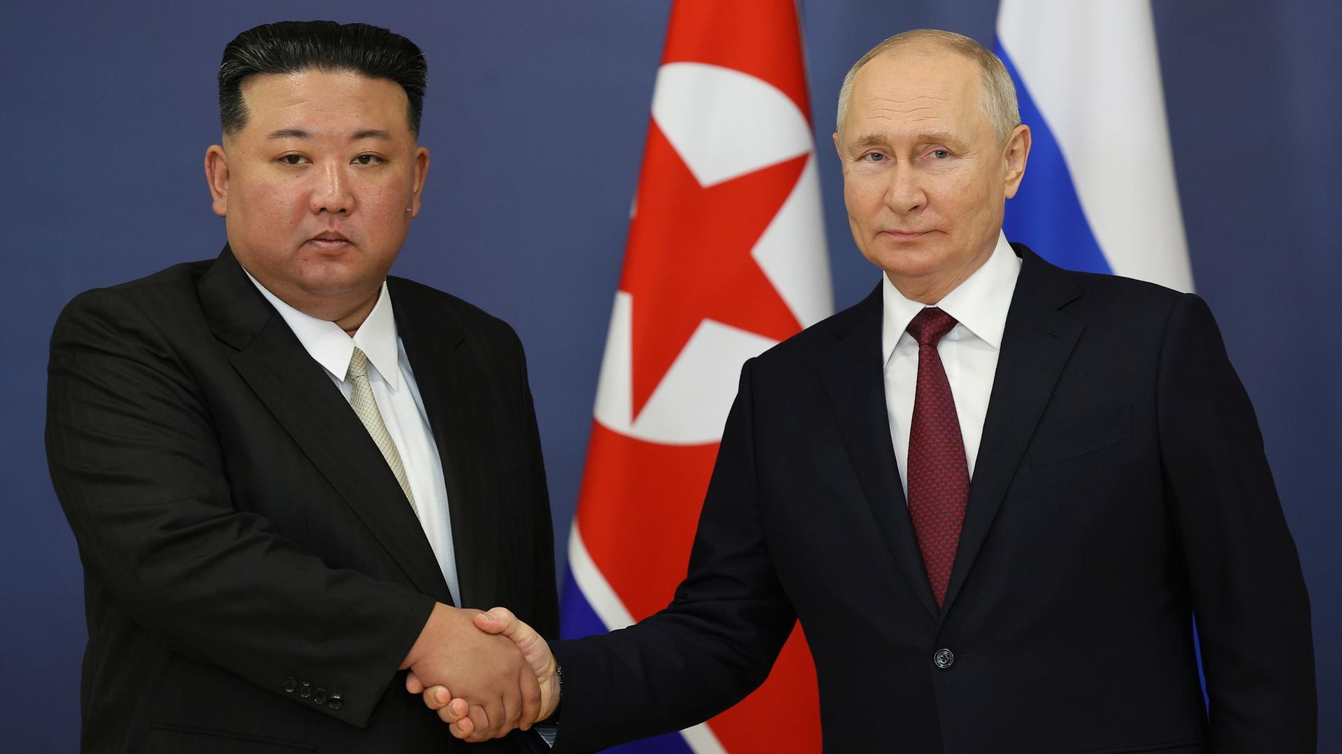 Links steht der Diktator von Nord-Korea, Kim Jong Un. Rechts steht der russische Präsident Wladimir Putin. Die beiden Männer geben sich die Hände.
