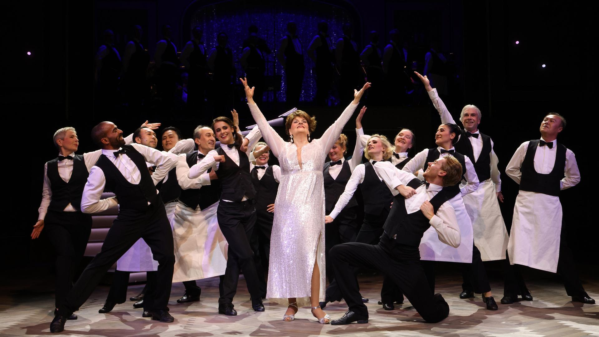 Eine Frau in einem weißen Kleid steht auf einer Bühne und hebt die Hände zum Himmel. Um sie herum zahlreiche Männer.