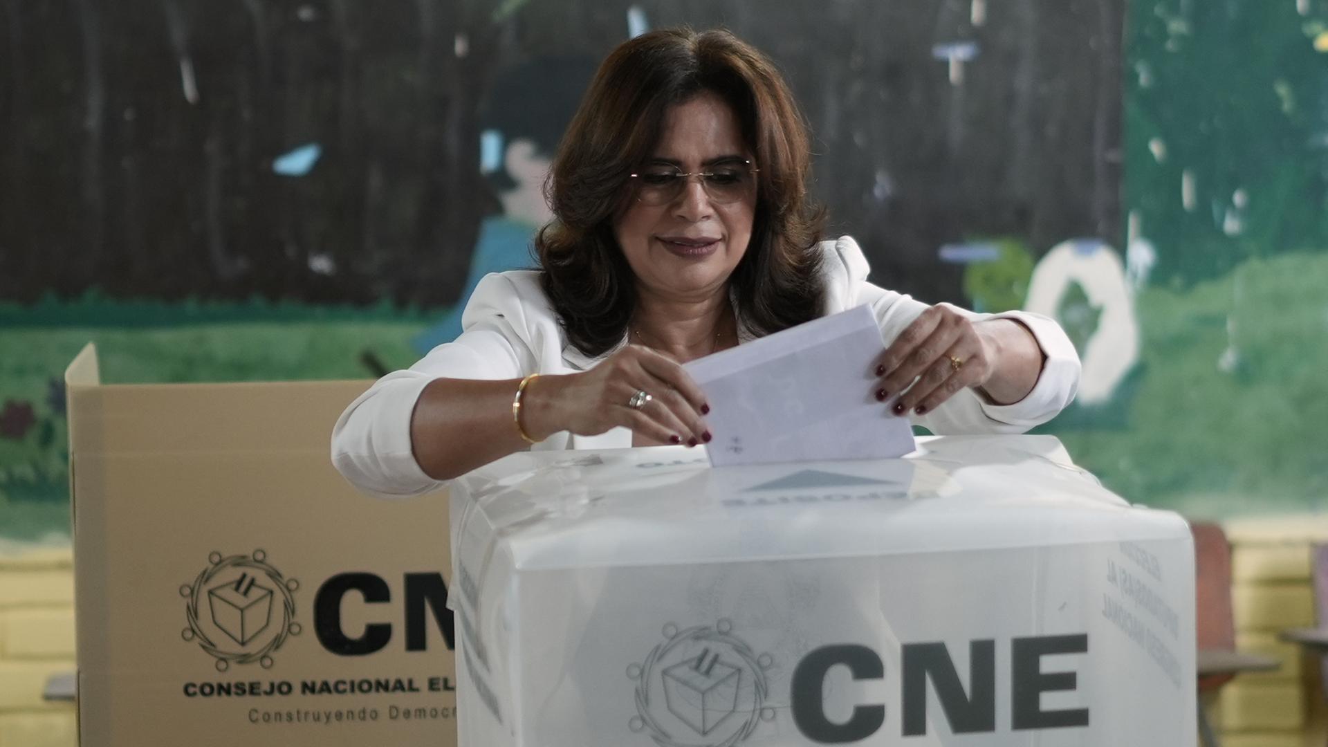 Moncada im weißen Hemd, mit langen Haaren und Brille steht hinter einer Wahlurne und steckt ihren Stimmzettel hinein. Dahinter ein Sichtschutz zur Stimmabgabe 