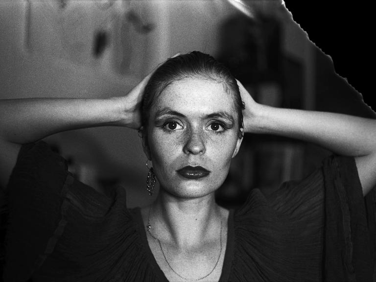 Aus der Serie "Lange Weile, 1983-1989" von der Fotografin Tina Bara. Eine Frau faltet die Hände hinter ihrem Kopf zusammen und blickt direkt in die Kamera.