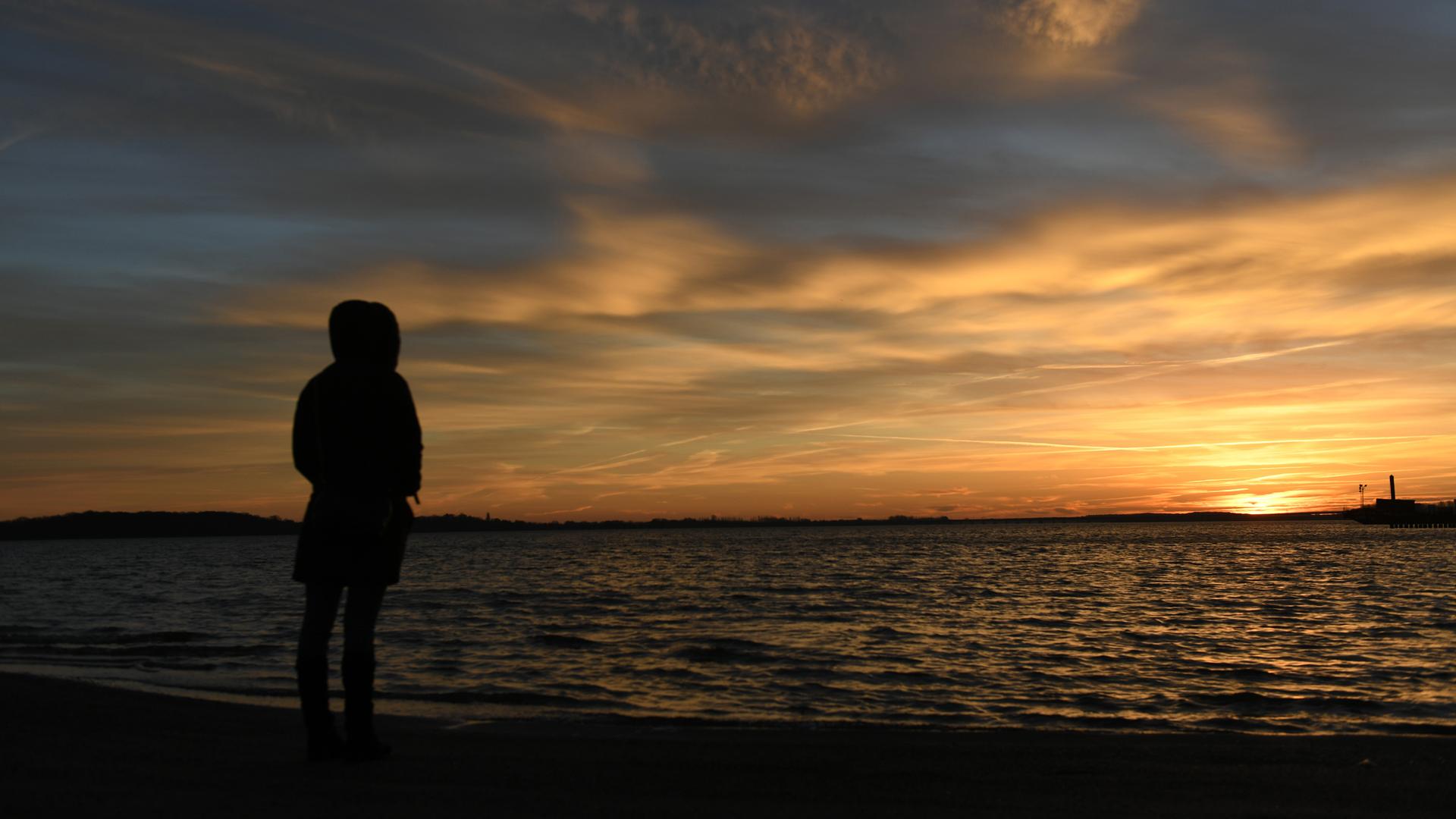 Eine Frau steht bei Sonnenaufgang am Strandbad von Stralsund und schaut auf das Wasser.