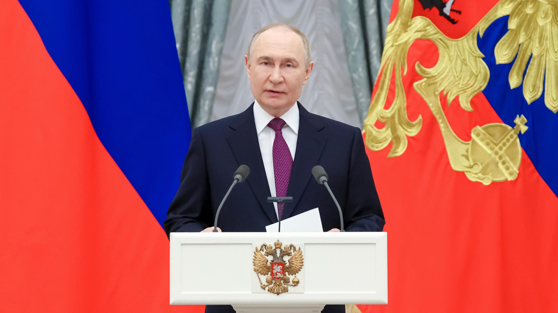 Der russische Präsident Wladimir Putin steht im dunklen Anzug an einem weißen Rednerpult. Im Hintergrund: Die Flagge Russlands. Der russische Präsident Wladimir Putin steht im dunklen Anzug an einem weißen Rednerpult. Im Hintergrund: Die Flagge Russlands.