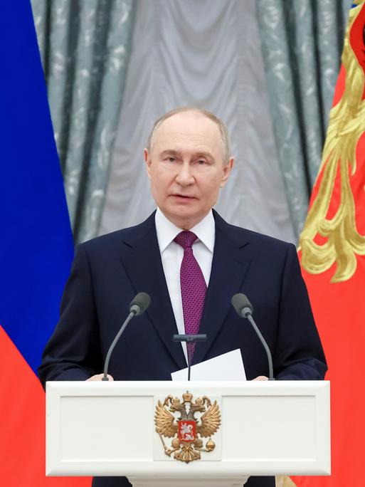 Der russische Präsident Wladimir Putin steht im dunklen Anzug an einem weißen Rednerpult. Im Hintergrund: Die Flagge Russlands.  