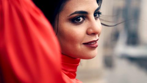 Fatma Said steht in einem tiefroten Kleid an einer Wand, während der Wind eine dunkle Haarsträhne aus ihrer Frisur gelöst hat.