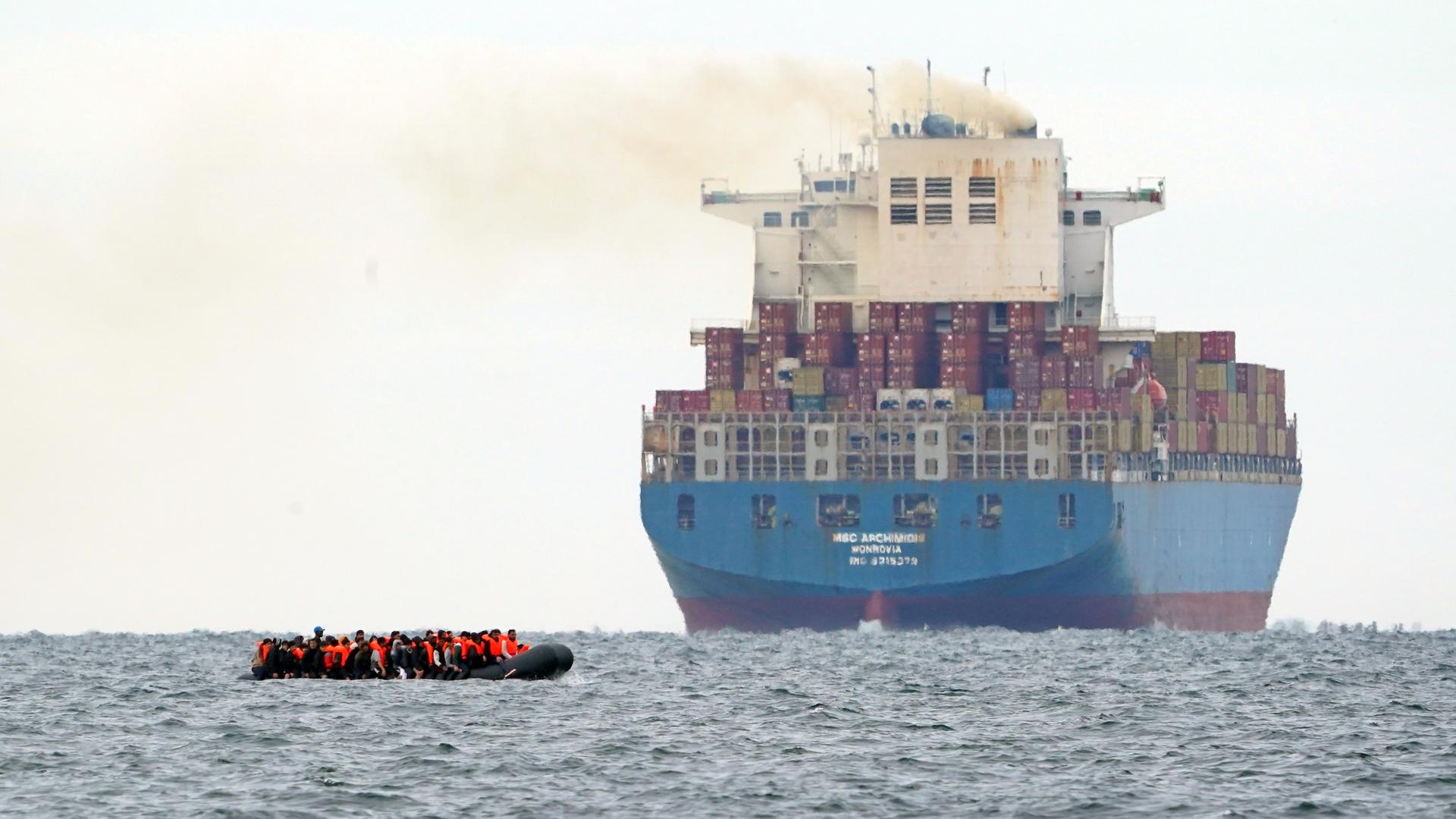 Flüchtlinge in einem viel zu kleinen Boot überqueren den Ärmelkanal von Frankreich nach England. Im Hintergrund ist ein Containerschiff zu sehen.