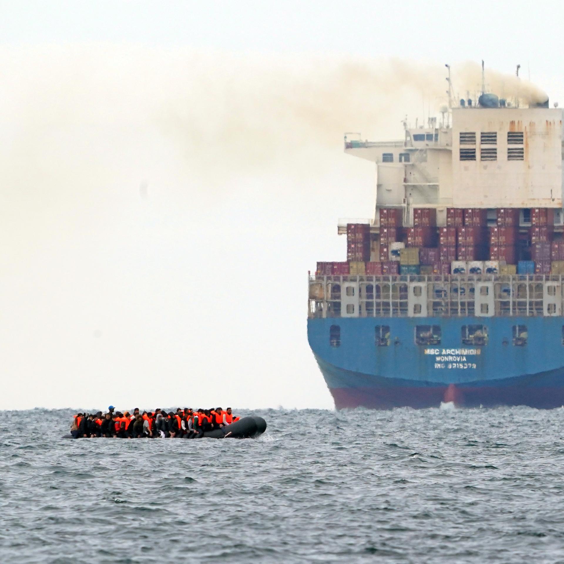 Flüchtlinge in einem viel zu kleinen Boot überqueren den Ärmelkanal von Frankreich nach England. Im Hintergrund ist ein Containerschiff zu sehen, 2023.