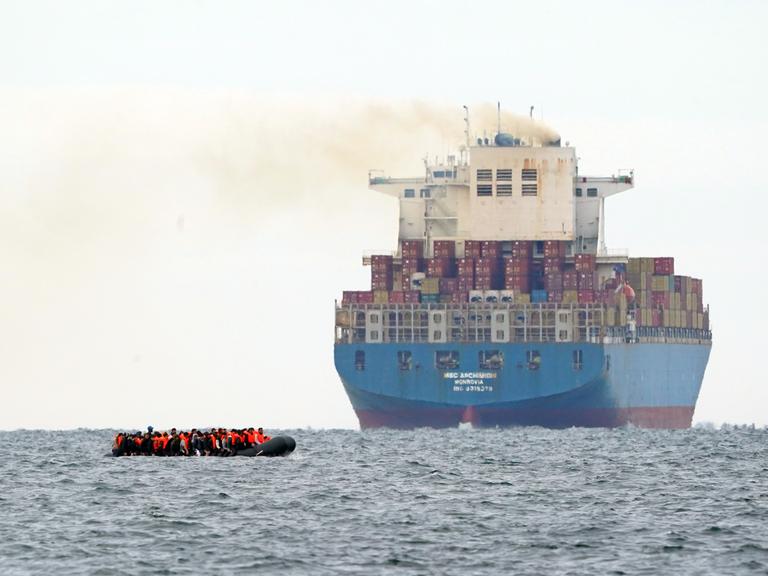 Flüchtlinge überqueren auf einem Schlauchboot den Ärmelkanal. Direkt daneben ist ein im Vergleich dazu riesig wirkendes Frachtschiff zu sehen.