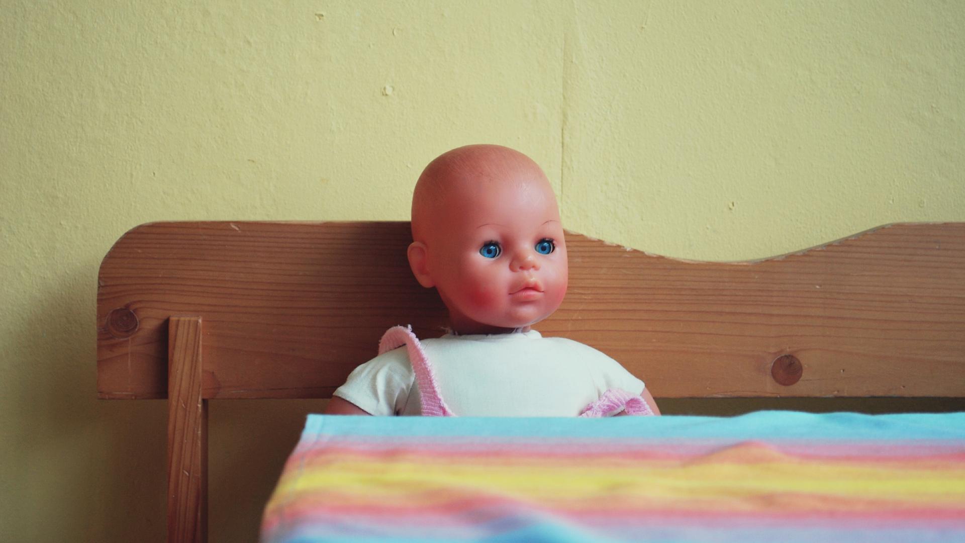Eine Puppe sitzt auf einer Holzbank in der Küche vor einem Tisch mit einer bunten Plastikdecke darauf.