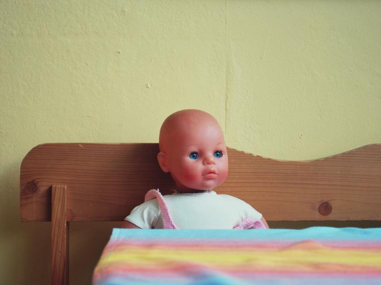Eine Puppe sitzt auf einer Holzbank in der Küche vor einem Tisch mit einer bunten Plastikdecke darauf.