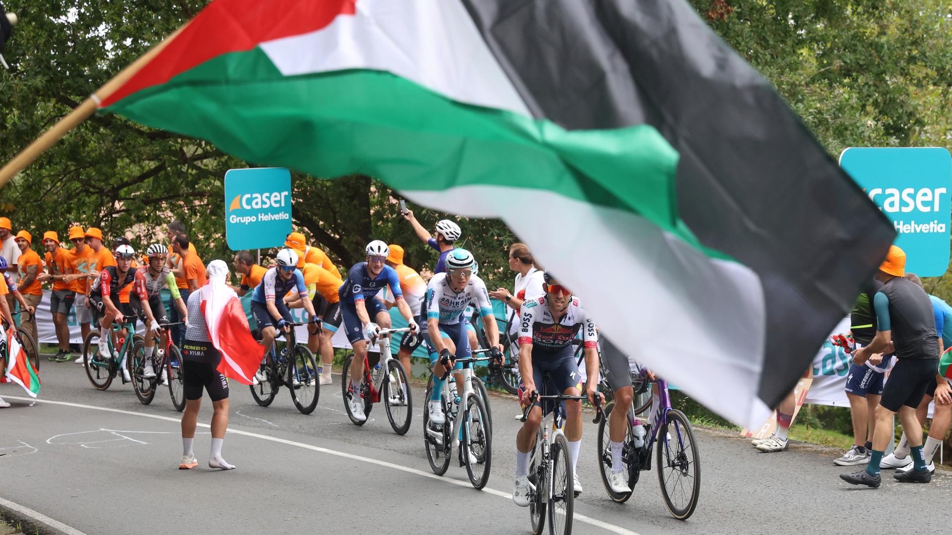 Fahrer der Spanien-Rundfahrt verschwinden fast hinter einer großen Palästina-Flagge