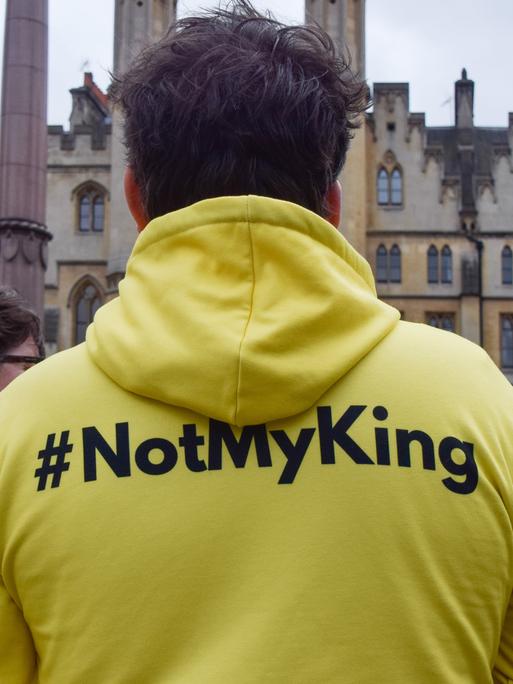 Eine Person, die einen neongelben Pullover mit der Aufschrift "#notmyking" trägt steht zusammen mit anderen Personenn vor einem Gebäude in London