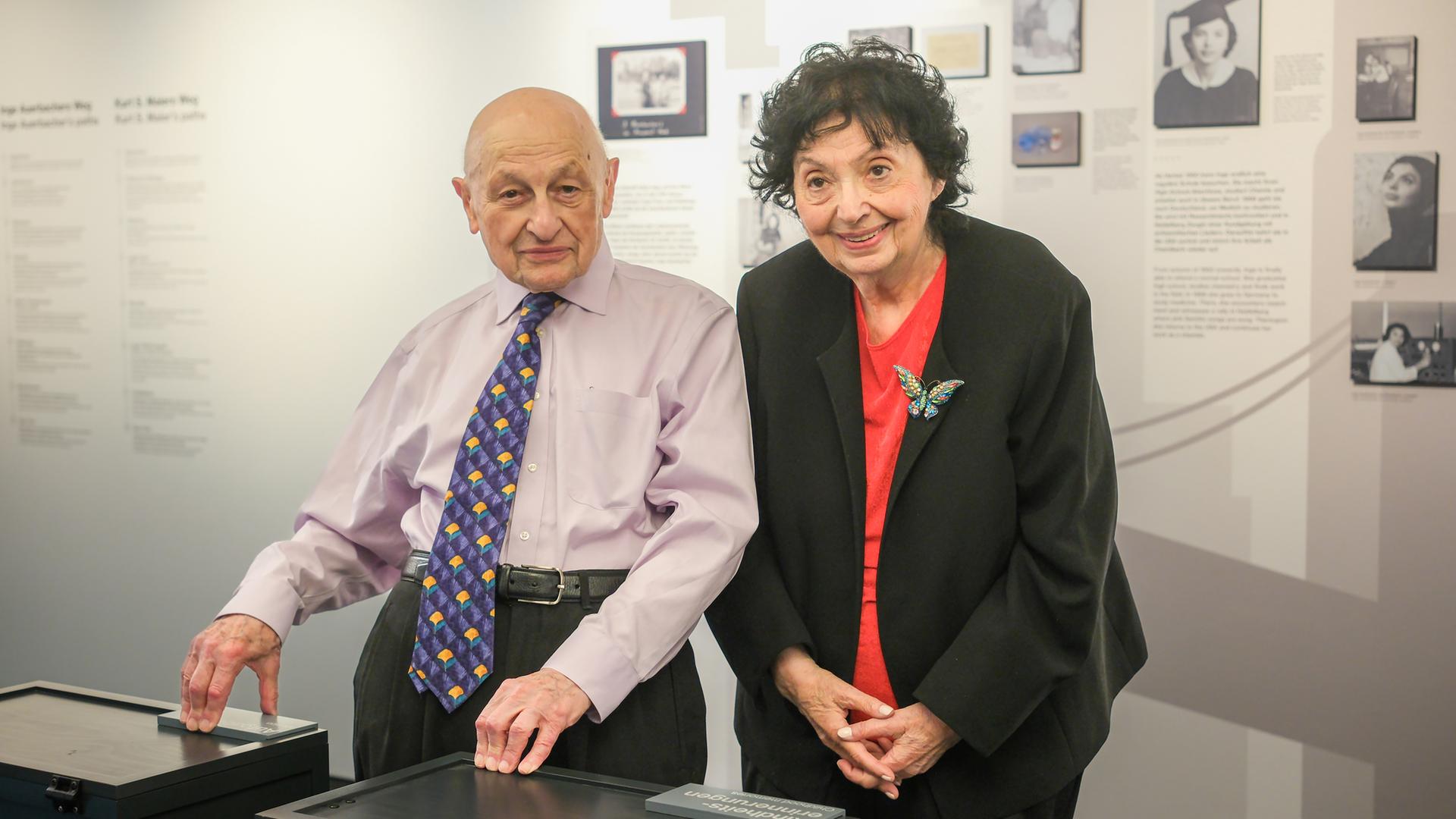 Kurt S. Maier und Inge Auerbacher stehen vor einer Ausstellungswand im Museum und lachen in die Kamera. Er trägt eine farbenfrohe Krawatte, sie eine Schmetterlingsbrosche an der Jacke. 
