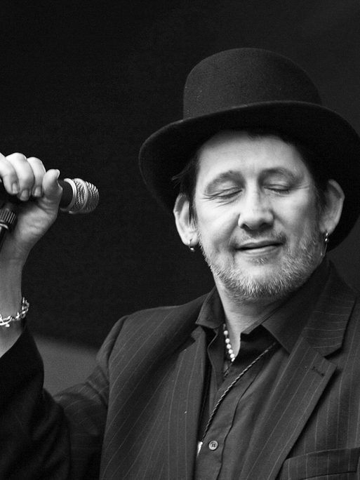 Schwarzweißfoto von Shane McGowan bei einem Konzert. Er steht versonnen lächelnd am Mikrofon.
