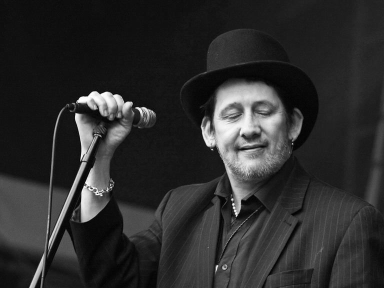 Schwarzweißfoto von Shane McGowan bei einem Konzert. Er steht versonnen lächelnd am Mikrofon.