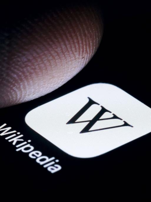 Die App von Wikipedia wird auf einem Smartphone angezeig, darüber ein Schatten eines Fingers.