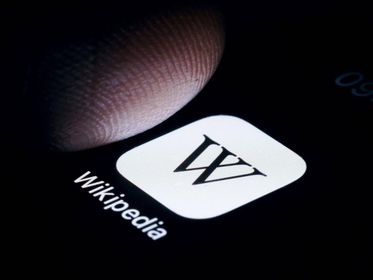 Die App von Wikipedia wird auf einem Smartphone angezeig, darüber ein Schatten eines Fingers.