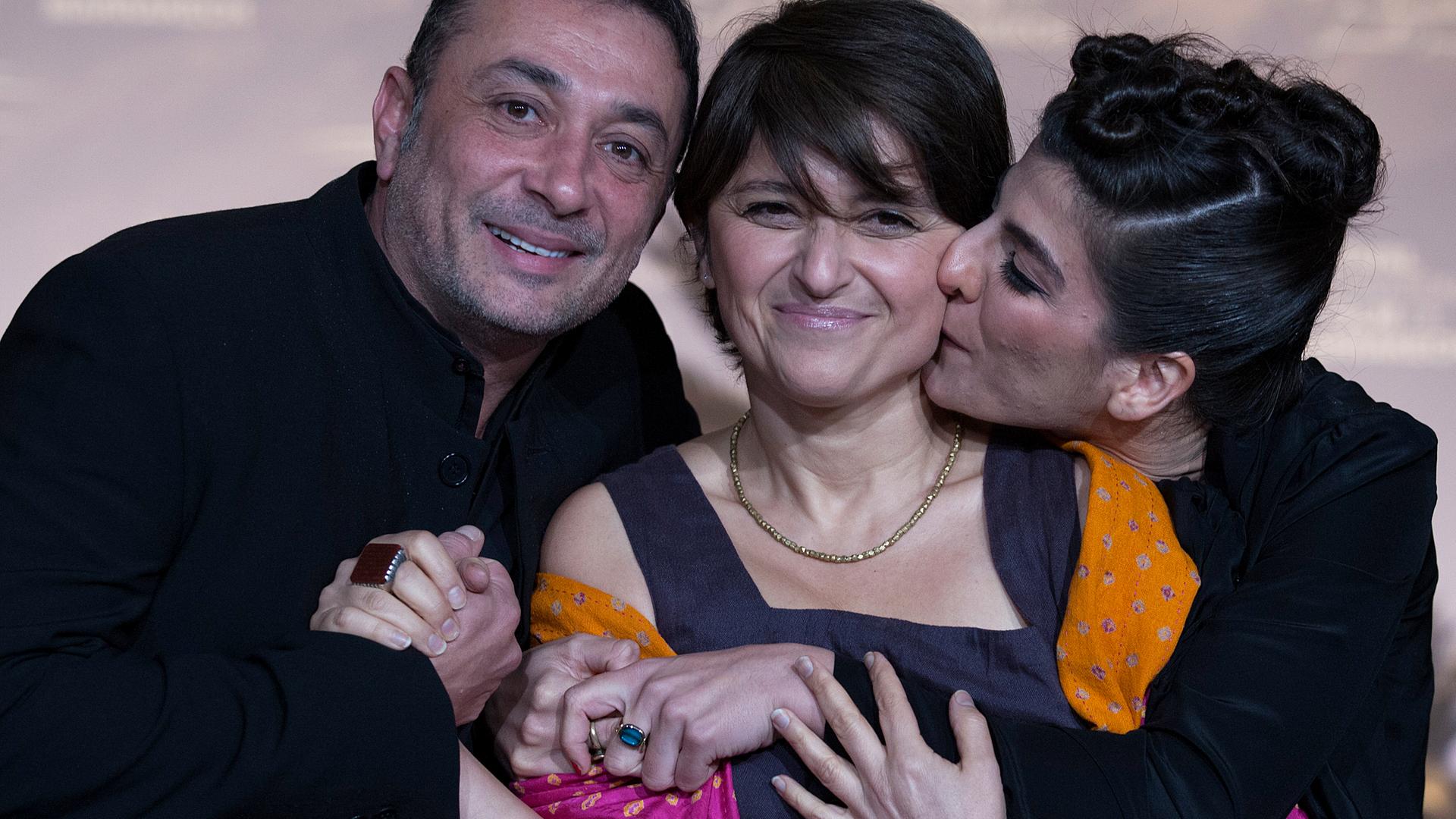 Sepideh Farsi steht zwischen einem Mann und einer Frau, die sie umarmen. Sie selbst lächelt in die Kamera.