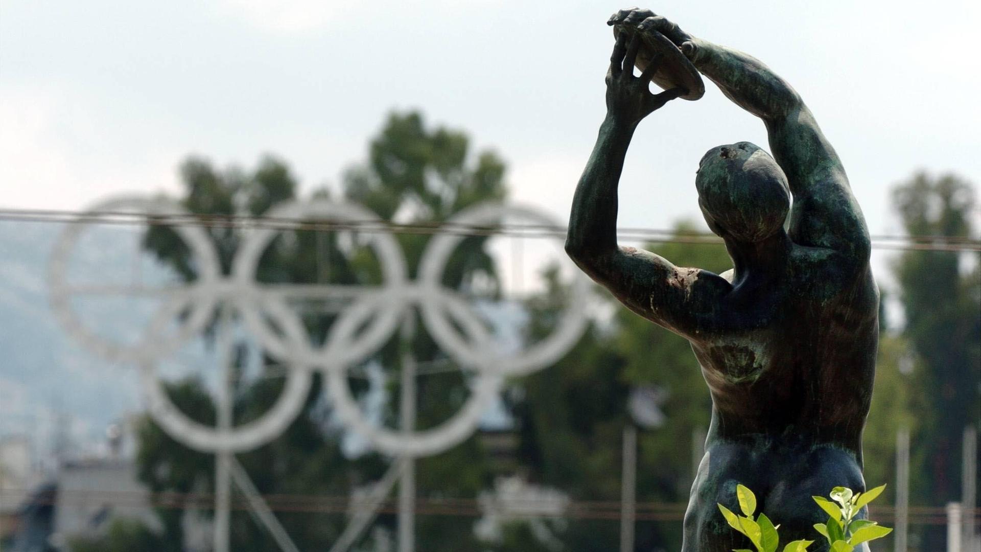 Eine typische griechische Statue eines Diskuswerfers ist im Vordergrund zu sehen. Im Bildhintergrund sind die fünf olympischen Ringe zu erkennen. Die Statue steht vor dem antike Olympiastadion in Athen.