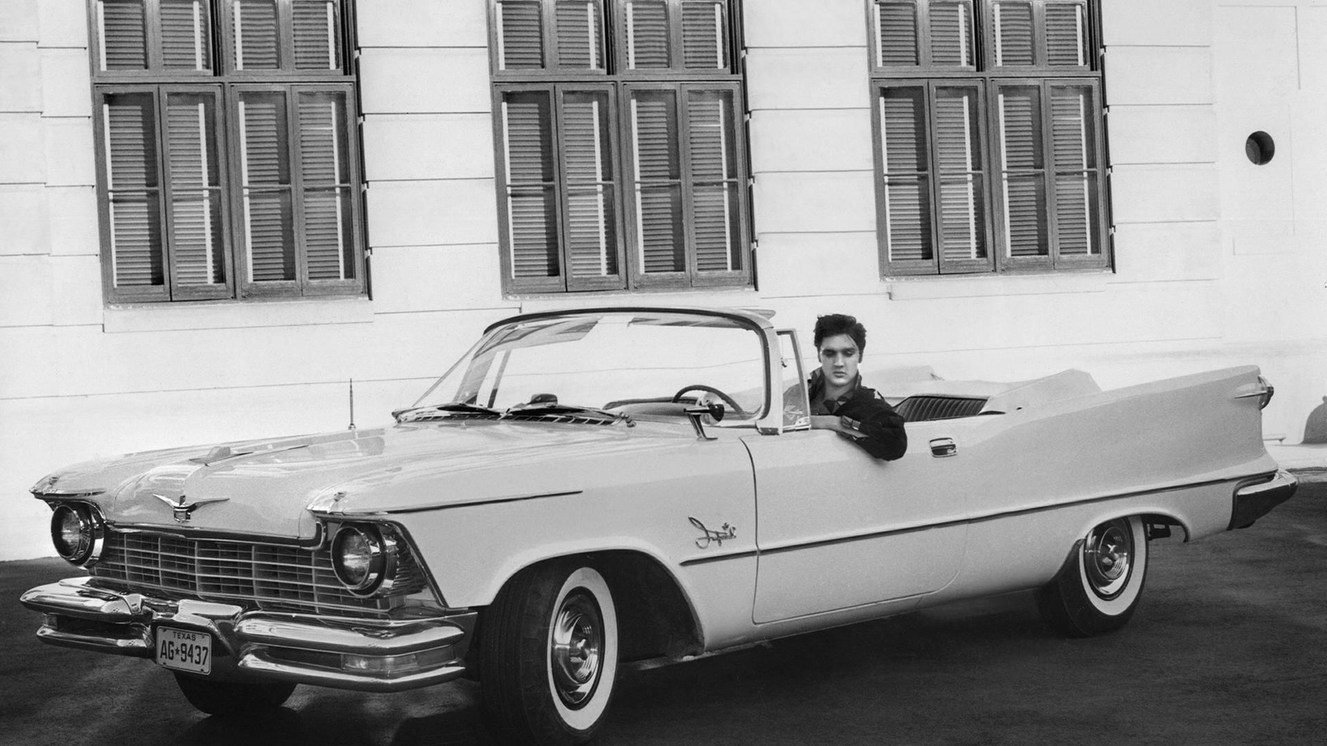 Der Sänger Elvis Presley in seinem Chrysler Imperial auf dem Studiogelände der Filmproduktionsfirma MGM in Hollywood, USA (1958). Sein Auto ist ein großes, weißes Cabriolet, das Verdeck ist geöffnet.