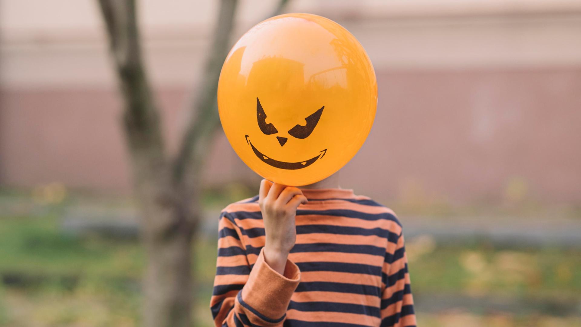 Ein Kind hält einen orangenen Luftballon mit Teufelsfratze vor sein Gesicht Ein Kind hält einen orangenen Luftballon mit Teufelsfratze vor sein Gesicht