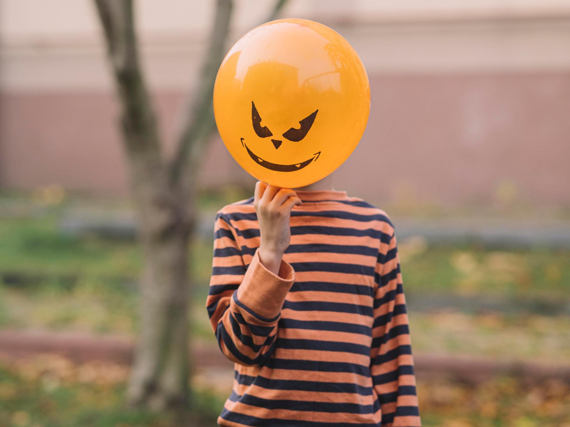 Ein Kind hält einen orangenen Luftballon mit Teufelsfratze vor sein Gesicht
