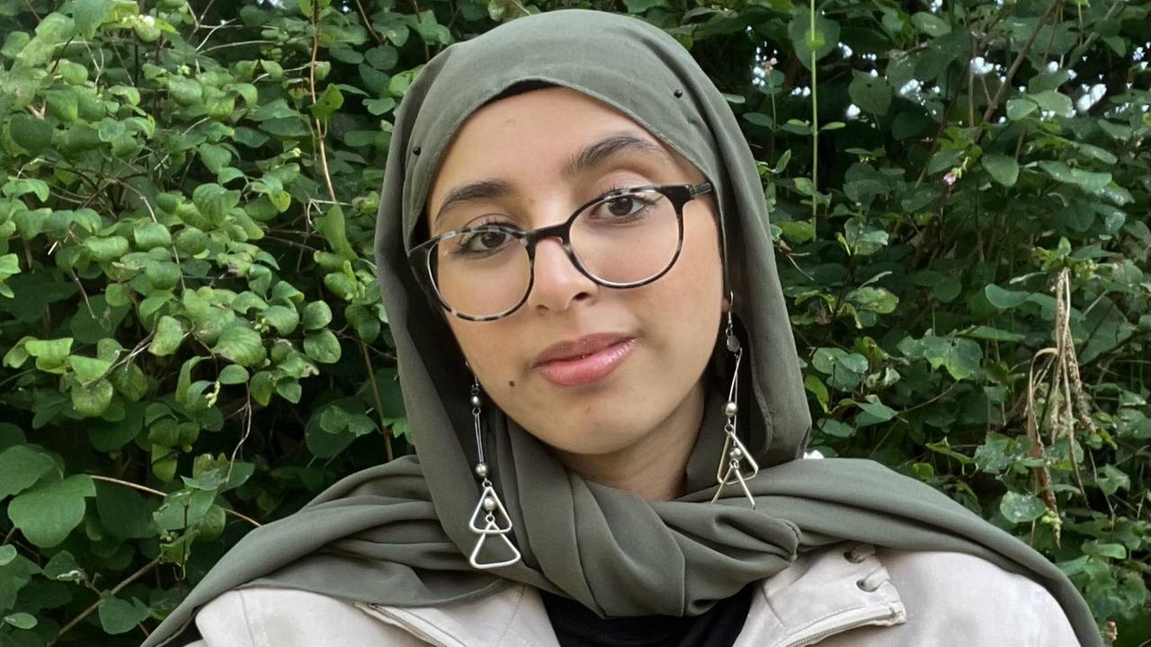 Tayiba schaut direkt in die Kamera. Sie trägt eine Brille mit schwarzem Rand und ein oliv-farbenes Kopftuch.