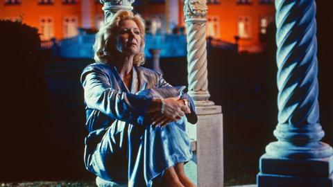 Christiane Hörbiger in einer Szene in der Folge "Der letzte Strohhalm" aus der Fernsehserie "Das Erbe der Guldenburgs". Sie hockt an eine Säule gelehnt vor einer Art Villa im Abendlicht.