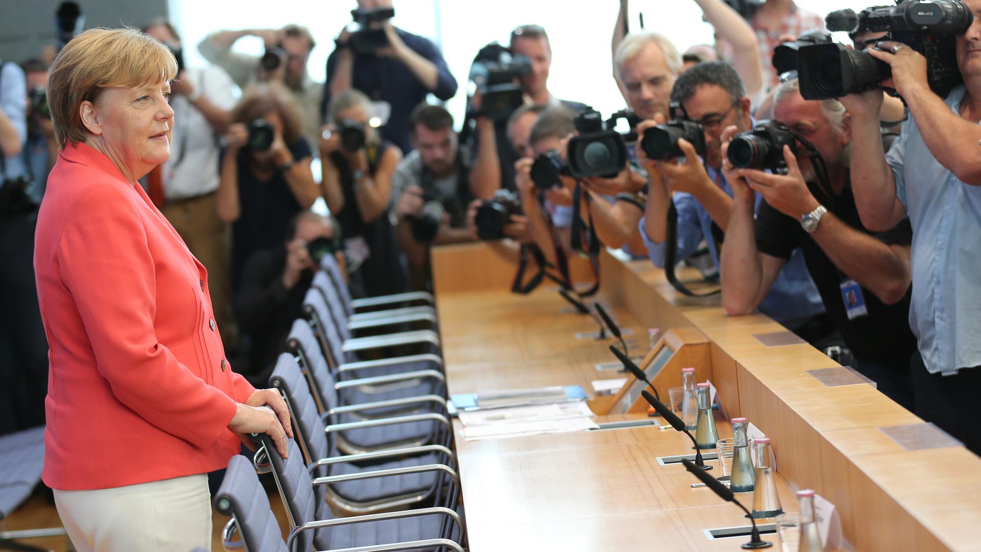 Angela Merkel steht im hellroten Blazer im Raum der Bundespressekonferenz in Berlin und wird von zahlreichen Fotografen fotografiert und gefilmt. 
