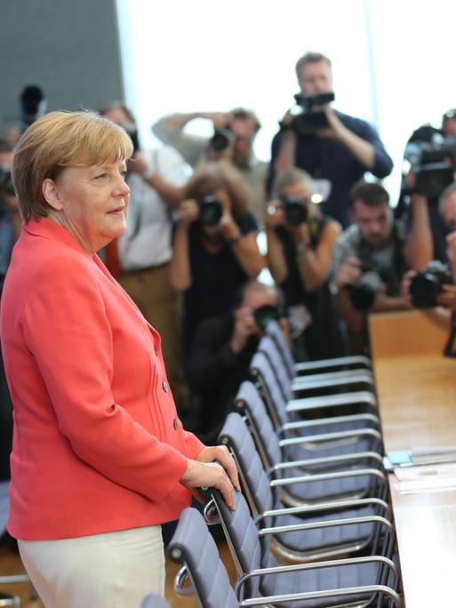 Angela Merkel steht im hellroten Blazer im Raum der Bundespressekonferenz in Berlin und wird von zahlreichen Fotografen fotografiert und gefilmt. 