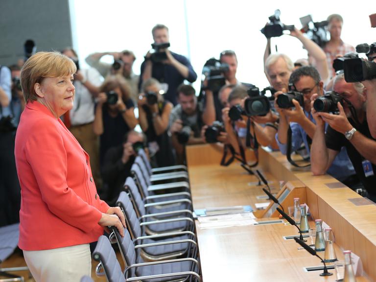 Angela Merkel steht im hellroten Blazer im Raum der Bundespressekonferenz in Berlin und wird von zahlreichen Fotografen fotografiert und gefilmt. 