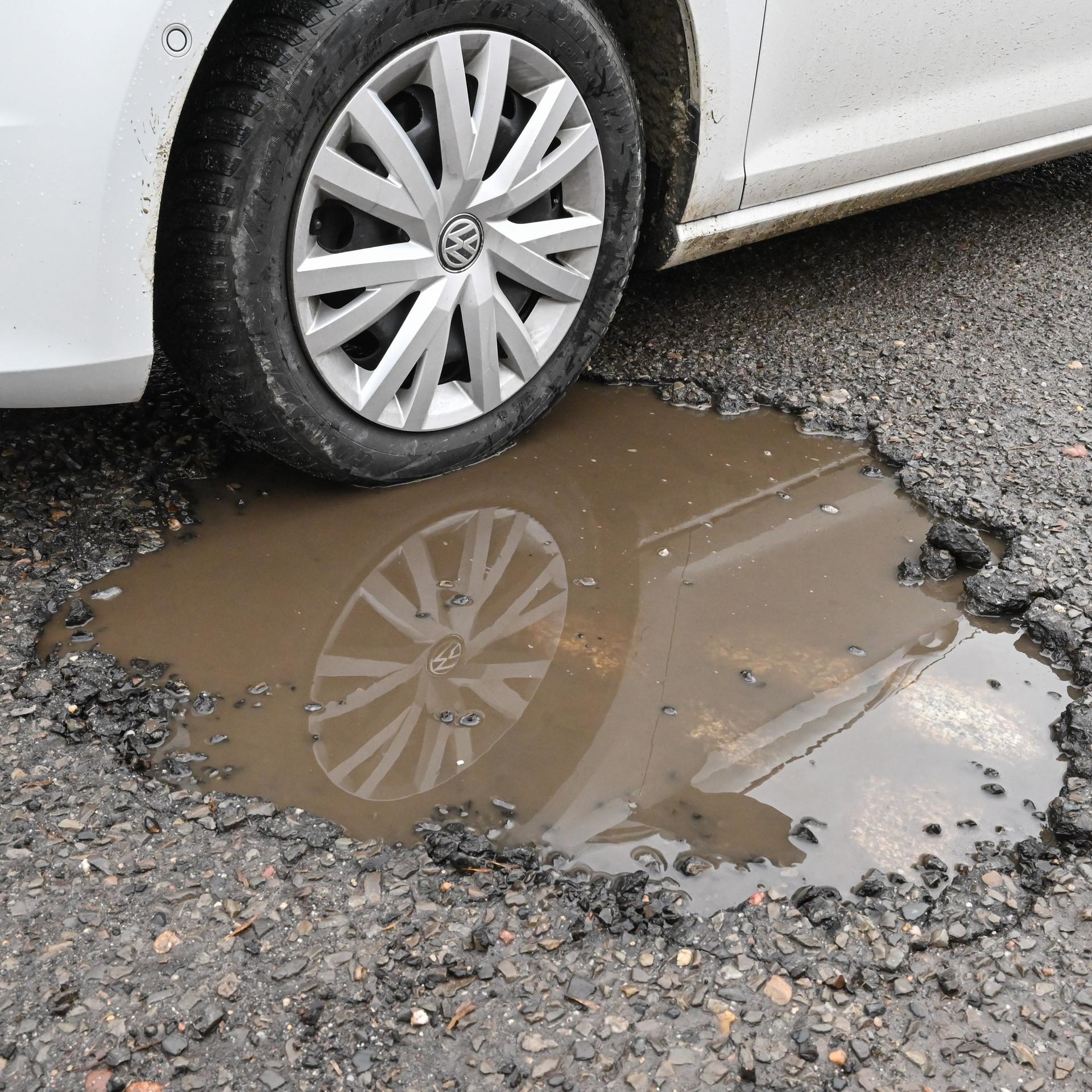Ein Pkw steht mit dem Vorderrad in einem mit Regenwasser gefüllten Schlagloch auf einer Straße. 