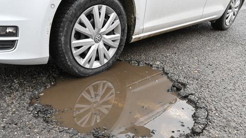 Ein Pkw steht mit dem Vorderrad in einem mit Regenwasser gefüllten Schlagloch auf einer Straße. 