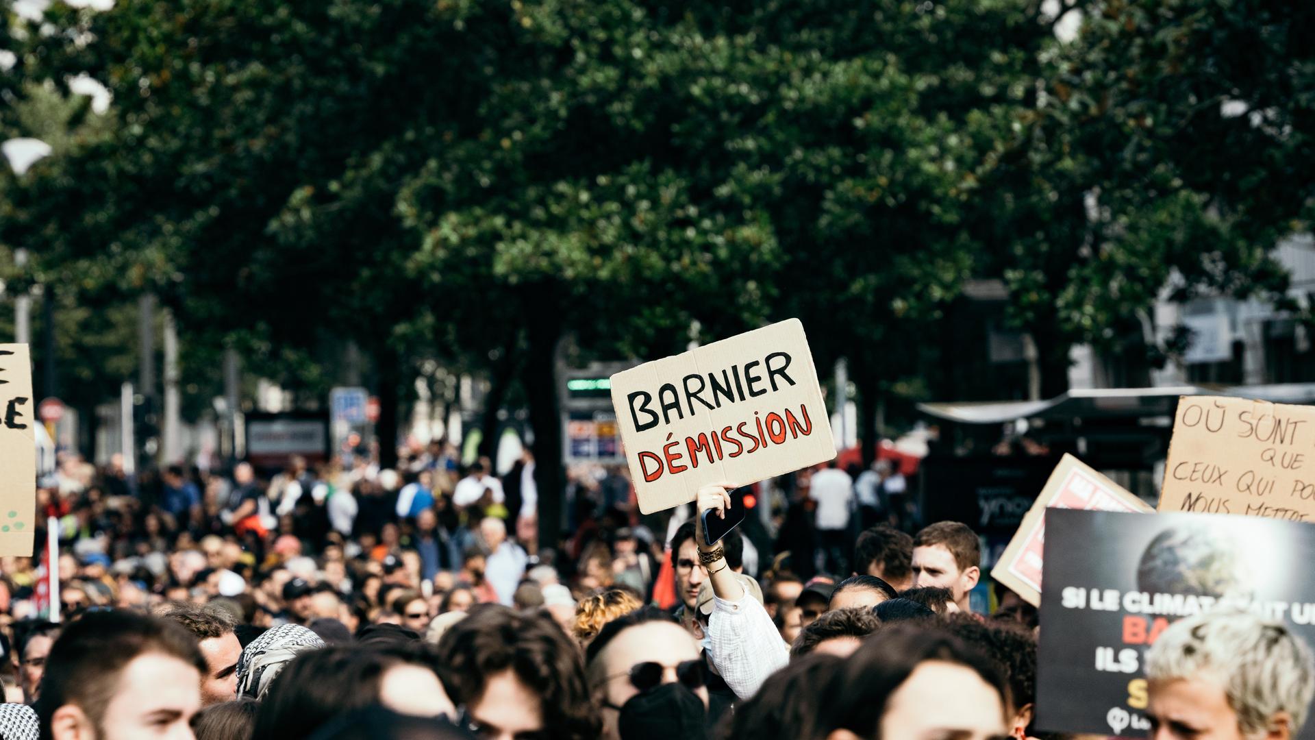 Demonstrierende in Frankreich. Aus der Mitte der Menschen wird ein Plakat hoch gehalten auf dem steht "Barnier Démission".