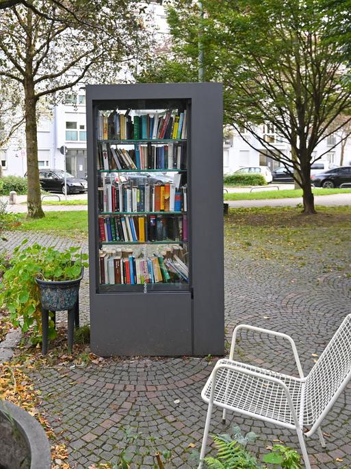 Öffentlicher Buecherschrank in München. Rechts davon eine weiße Bank und links ein Strauch mit Blüten.