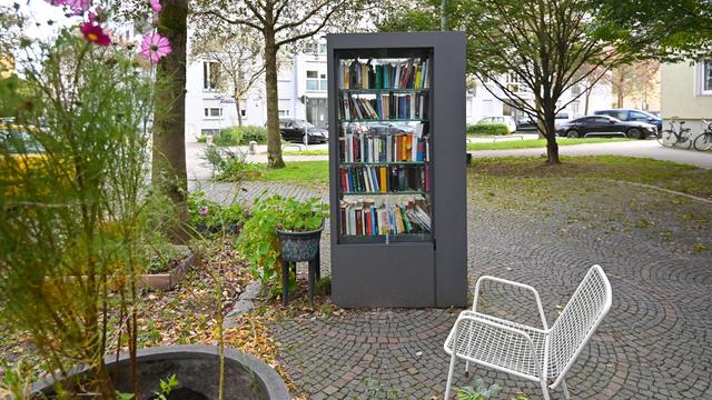 Öffentlicher Buecherschrank in München. Rechts davon eine weiße Bank und links ein Strauch mit Blüten.