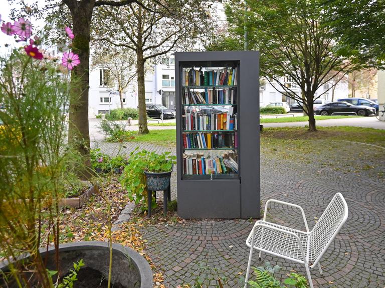 Öffentlicher Buecherschrank in München. Rechts davon eine weiße Bank und links ein Strauch mit Blüten.