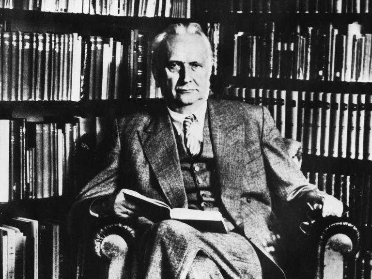 Karl Jaspers sitzt auf einem Lesesessel vor einem Bücherregal und blickt ernst in die Kamera